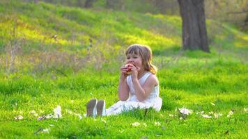 Happy little girl is sitting on the green grass and eating an apple. 4K video