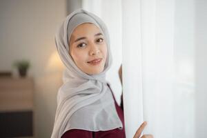 Muslim Islam woman wearing hijab or grey scarf stands in front of a window. She is smiling and looking at the camera photo