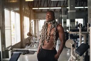 A man wearing a chain around his neck stands in a gym. The man is in a serious or determined mood, possibly preparing for a workout or a competition photo