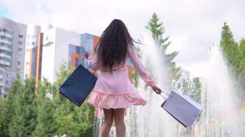 Beautiful girl near a fountain in the park after shopping with packages in hands. slow motion. video