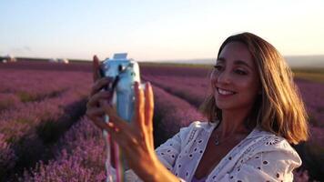 Smiling happy woman photographing with instant camera in lavender field on sunny day. Young woman shooting with blue instant camera - Trendy girl taking selfie outdoor video
