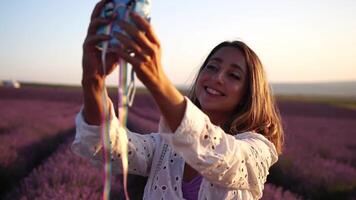 glimlachen gelukkig vrouw fotograferen met ogenblik camera in lavendel veld- Aan zonnig dag. jong vrouw het schieten met blauw ogenblik camera - modieus meisje nemen selfie buitenshuis video