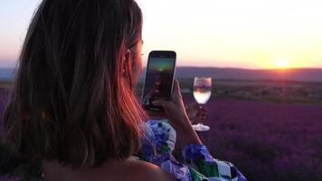 femme dans lavande champ. branché fille prise selfie Extérieur avec une verre de Champagne dans lavande champ sur été le coucher du soleil. souriant content femme photographier avec téléphone intelligent. video