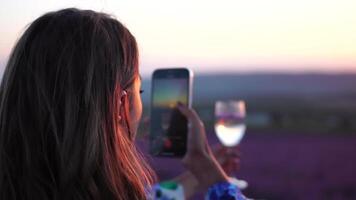 Woman in lavender field. Trendy girl taking selfie outdoor with a glass of champagne in lavender field on summer sunset. Smiling happy woman photographing with smartphone. video