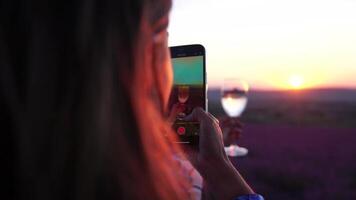 mujer en lavanda campo. de moda niña tomando selfie al aire libre con un vaso de champán en lavanda campo en verano puesta de sol. sonriente contento mujer fotografiando con teléfono inteligente video