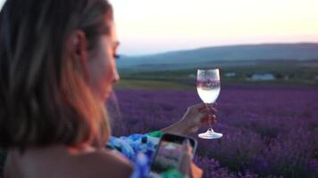 mulher dentro lavanda campo. na moda menina levando selfie ao ar livre com uma vidro do champanhe dentro lavanda campo em verão pôr do sol. sorridente feliz mulher fotografando com Smartphone. video