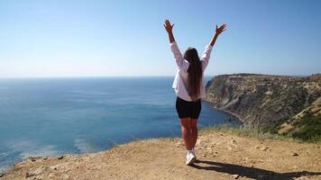 Woman summer travel sea. Happy carefree lady in a pink shirt and white skirt raises arms on sea ocean cliff. Sunny day, capturing triumph and admiration of breathtaking view, celebrating achievement. video