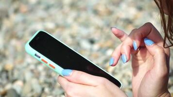 vrouw met smartphone. detailopname van vrouw handen Holding verticaal mobiel telefoon en vegen omhoog vinger toepassing bladzijde tegen achtergrond van zee en strand video