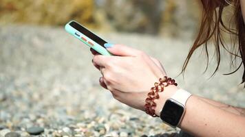 Woman with smartphone. Close-up of woman's hands holding vertical mobile phone and swiping up finger application page against background of sea and beach video