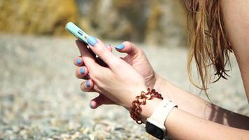 Woman with smartphone. Close-up of woman's hands holding vertical mobile phone and swiping up finger application page against background of sea and beach video