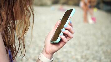 Woman with smartphone. Close-up of woman's hands holding vertical mobile phone and swiping up finger application page against background of sea and beach video