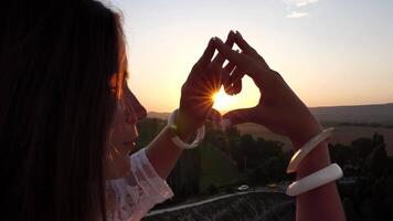 contento mujer en blanco boho vestir haciendo corazón firmar con manos en puesta de sol en montañas. romántico mujer con largo pelo en pie con su espalda en el puesta de sol en naturaleza en verano con abierto manos. video