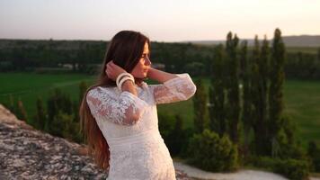 Lycklig kvinna i vit boho klänning på solnedgång i berg. romantisk kvinna med lång hår stående med henne tillbaka på de solnedgång i natur i sommar med öppen händer. silhuett. natur. solnedgång. video