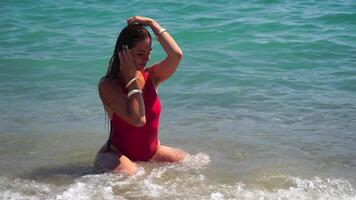 Jeune femme avec longue cheveux dans une rouge maillot de bain et bracelets dans boho style profiter le vagues sur le plage dans été. adorable content féminin relaxant à été plage ayant vacances. lent mouvement. video