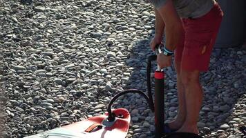 Man Inflates Standup Puddle Board. Person pumping on beach close up shot of pump and person hands in focus while constantly pumping. Slow motion video