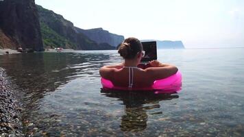 Frau Freiberufler funktioniert auf Laptop Schwimmen im Meer auf Rosa aufblasbar Ring. glücklich Tourist im Sonnenbrille schwebend auf aufblasbar Krapfen und Arbeiten auf Laptop Computer im Ruhe Ozean. Fernbedienung Arbeiten irgendwo video