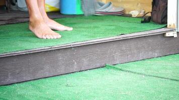 unrecognizable man sweeping the green carpet of artificial lawn on the beach from pebbles with broom. video