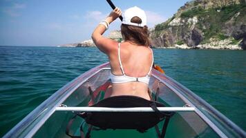 Young attractive brunette woman with long hair in white swimsuit, swimming on transparent kayak around volcanic rocks, like in Iceland. Back view. Summer holiday vacation and travel concept. video