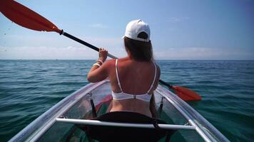 Young attractive brunette woman with long hair in white swimsuit, swimming on transparent kayak around volcanic rocks, like in Iceland. Back view. Summer holiday vacation and travel concept. video