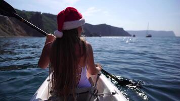 Young attractive brunette woman with long hair in white swimsuit, swimming on kayak around volcanic rocks, like in Iceland. Back view. Christmas holiday vacation and travel concept. video