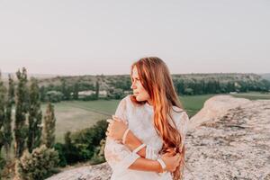 contento mujer en blanco boho vestir en puesta de sol en montañas. romántico mujer con largo pelo en pie con su espalda en el puesta de sol en naturaleza en verano con abierto manos. silueta. naturaleza. puesta de sol. foto