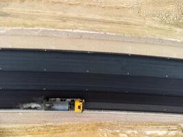 yellow truck pouring bitumen over a new road. Aerial drone shot. Road construction in progress on slope nature canyon. Infrastructure development and logistics. photo