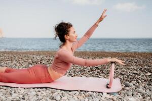 Woman sea pilates. Sporty happy middle aged woman practicing fitness on beach near sea, smiling active female training with ring on yoga mat outside, enjoying healthy lifestyle, harmony and meditation photo