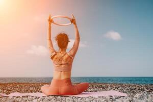 mujer mar pilates deportivo contento medio Envejecido mujer practicando aptitud en playa cerca mar, sonriente activo hembra formación con anillo en yoga estera afuera, disfrutando sano estilo de vida, armonía y meditación foto