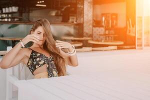 contento mujer retrato en cafetería. boho elegante Moda estilo. al aire libre foto de joven contento mujer con largo cabello, soleado clima al aire libre sentado en moderno cafetería.