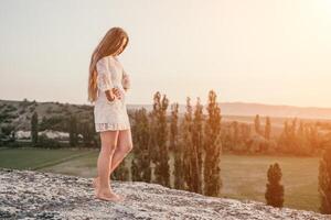 Happy woman in white boho dress on sunset in mountains. Romantic woman with long hair standing with her back on the sunset in nature in summer with open hands. Silhouette. Nature. Sunset. photo