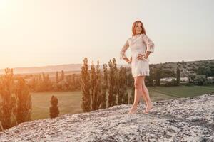 Happy woman in white boho dress on sunset in mountains. Romantic woman with long hair standing with her back on the sunset in nature in summer with open hands. Silhouette. Nature. Sunset. photo