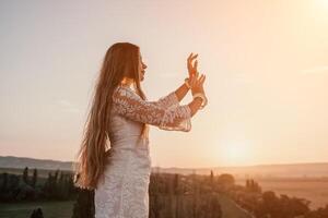 Happy woman in white boho dress on sunset in mountains. Romantic woman with long hair standing with her back on the sunset in nature in summer with open hands. Silhouette. Nature. Sunset. photo