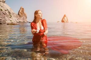 Woman travel sea. Happy tourist in red dress enjoy taking picture outdoors for memories. Woman traveler posing in sea beach, surrounded by volcanic mountains, sharing travel adventure journey photo