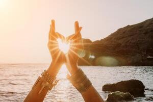 Woman sea yoga. Happy woman in a white swimsuit and boho-style bracelets making a heart shape with her hands while practicing yoga on a mat by sea at sunset. Healthy lifestyle, harmony and meditation photo