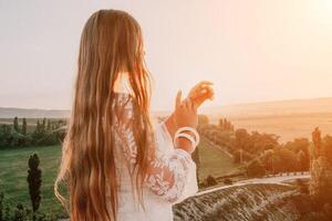 Happy woman in white boho dress on sunset in mountains. Romantic woman with long hair standing with her back on the sunset in nature in summer with open hands. Silhouette. Nature. Sunset. photo