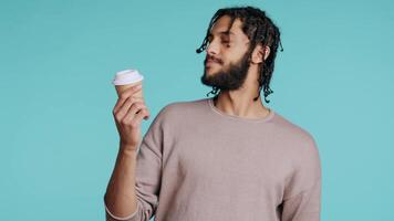 Man enjoys fresh coffee from disposable paper cup early in the morning to wake up and be energized. Person drinks hot beverage from brown recycled takeaway cappuccino cup, studio background, camera B video