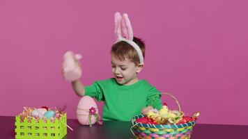 joyeux Jeune enfant en jouant autour avec de fête peint décorations, montrant une lapin jouet et une rose Oeuf dans de face de caméra. souriant petit garçon avec lapin oreilles ayant amusement avec ornements. caméra un. video