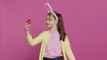 entusiasta piccolo bambino Filatura in giro nel davanti di telecamera, presentazione sua rosso dipinto uovo per Pasqua Domenica festa. allegro bambino piccolo con coniglietto orecchie volteggiare e fare un' piroetta. telecamera un. video