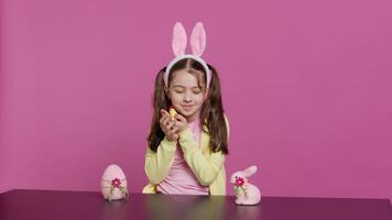 Joyful little girl playing with festive easter decorations in studio, creating arrangements with a chick, rabbit and egg. Smiling cute toddler with bunny ears showing colorful ornaments. Camera A. video