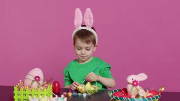 contento pequeño niño arreglando huevos y polluelo en un cesta en preparación para Pascua de Resurrección domingo celebraciones, creando festivo decoraciones en estudio. pequeño chico teniendo divertido colorante. cámara una. video