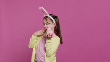 Jolly schoolgirl holding a cute rabbit toy and blowing kisses in studio, being adorable with festive decorations for easter holiday event. Happy child wearing bunny ears and pigtails. Camera A. video