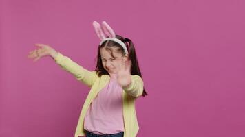 aanbiddelijk schattig kind zetten konijn oren en golvend Bij camera, genieten van Pasen zondag viering tegen roze achtergrond. glimlachen vrolijk schoolmeisje met vlechten gezegde Hallo. camera a. video