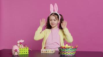 énergique Jeune fille avec adorable lapin oreilles agitant dans studio, en disant Bonjour et salutation Quelqu'un tandis que elle crée Pâques décorations. joyeux bambin posant contre rose toile de fond. caméra un. video