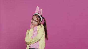 sonriente pequeño niña soñando acerca de regalos y comida para Pascua de Resurrección domingo celebracion, sensación emocionado acerca de primavera día festivo. joven niño con conejito orejas posando en contra rosado antecedentes. cámara una. video