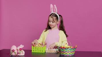 Joyful little girl showing her handcrafted festive basket on camera, creating a colorful handmade arrangement in time for easter holiday. Small toddler with bunny ears decorating eggs. Camera A. video