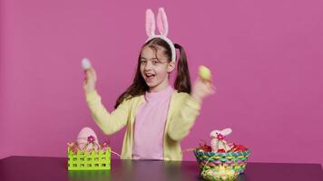 Enthusiastic young girl playing peek a boo in front of camera, using painted easter eggs against pink background. Joyful lovely toddler feeling excited about spring holiday festivity. Camera A. video