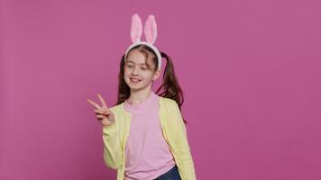 Young cheerful kid with pigtails showing peace sign in studio, feeling excited and confident about easter sunday celebration. Smiling youngster does signs against background. Camera A. video