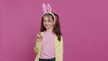 encantador dulce niña posando en estudio con dedos cruzado y esperando a recibir regalos para Pascua de Resurrección fiesta celebracion. alegre joven niño con conejito orejas preguntando para bueno suerte y fortuna. cámara una. video