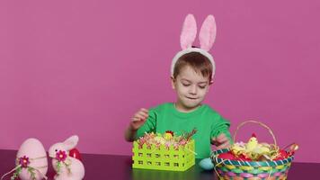 Sweet young boy making colorful arrangements for Easter holiday festivity, putting painted eggs in a handcrafted basket. Ecstatic joyful kid using crafting materials to create decorations. Camera A. video