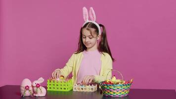 Smiling happy schoolgirl presenting a handmade decorated basket in studio, making easter holiday preparations against pink background. Young child showing handcrafted arrangements. Camera A. video